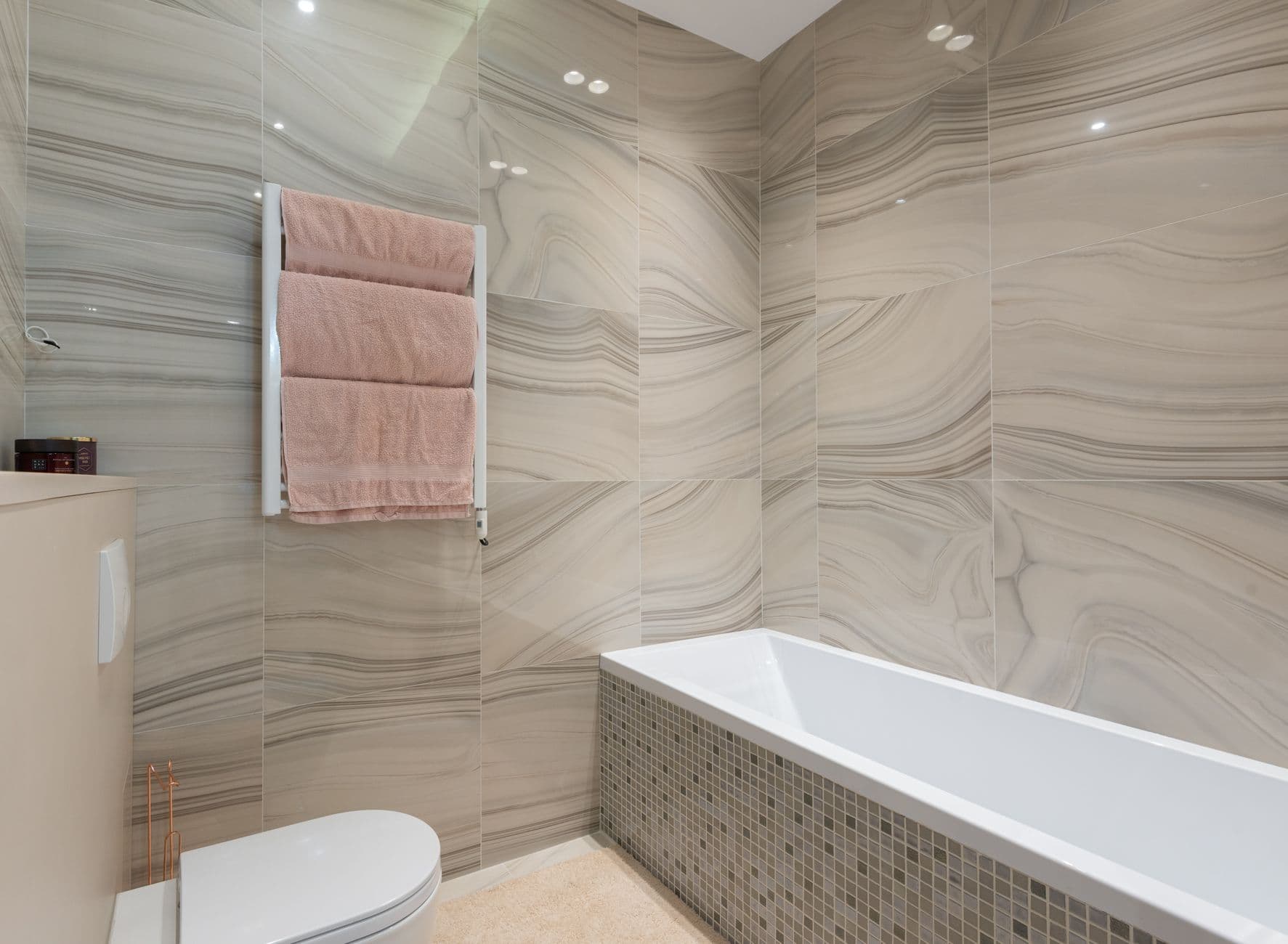 Pale stone bathroom with a bath, toilet and daylight across the wall finish
